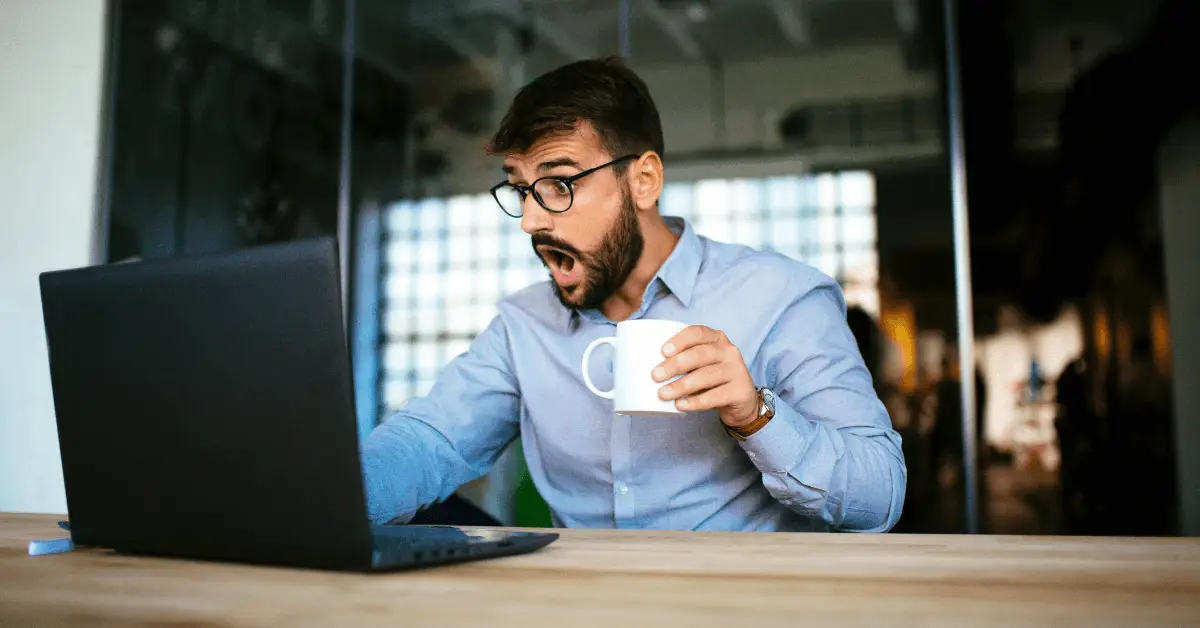 A person in glasses holding a cup of coffee and looking at a laptop.