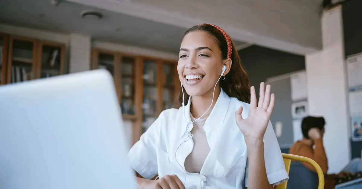 Image shows mixed race woman in a white shirt sitting in front of her laptop screen waving