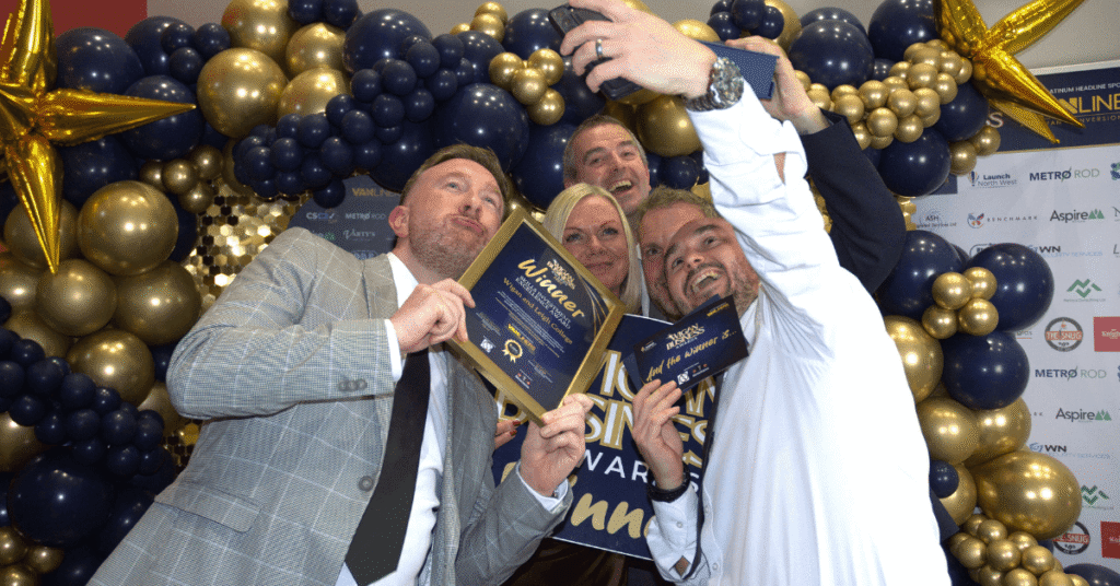 A group of people are posing for a selfie in front of a balloon arch and a backdrop with Wigan business award logos. They are holding awards and smiling.