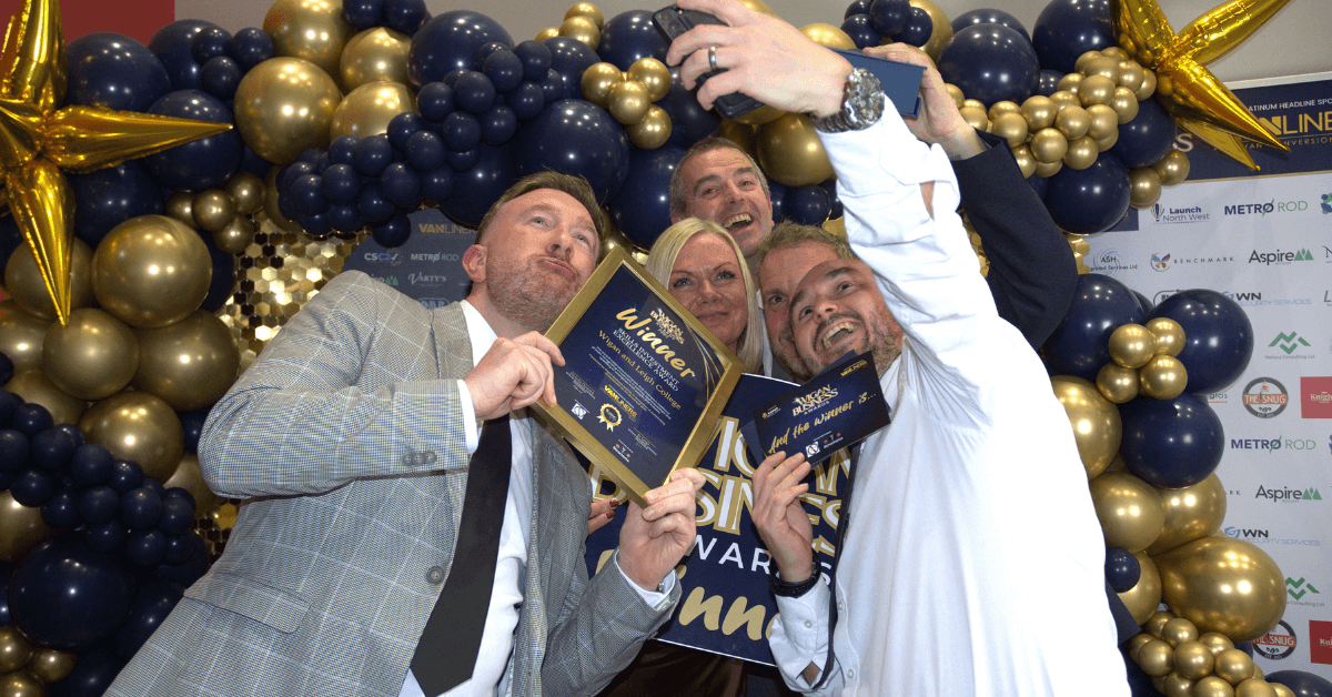 A group of people are posing for a selfie in front of a balloon arch and a backdrop with Wigan business award logos. They are holding awards and smiling.