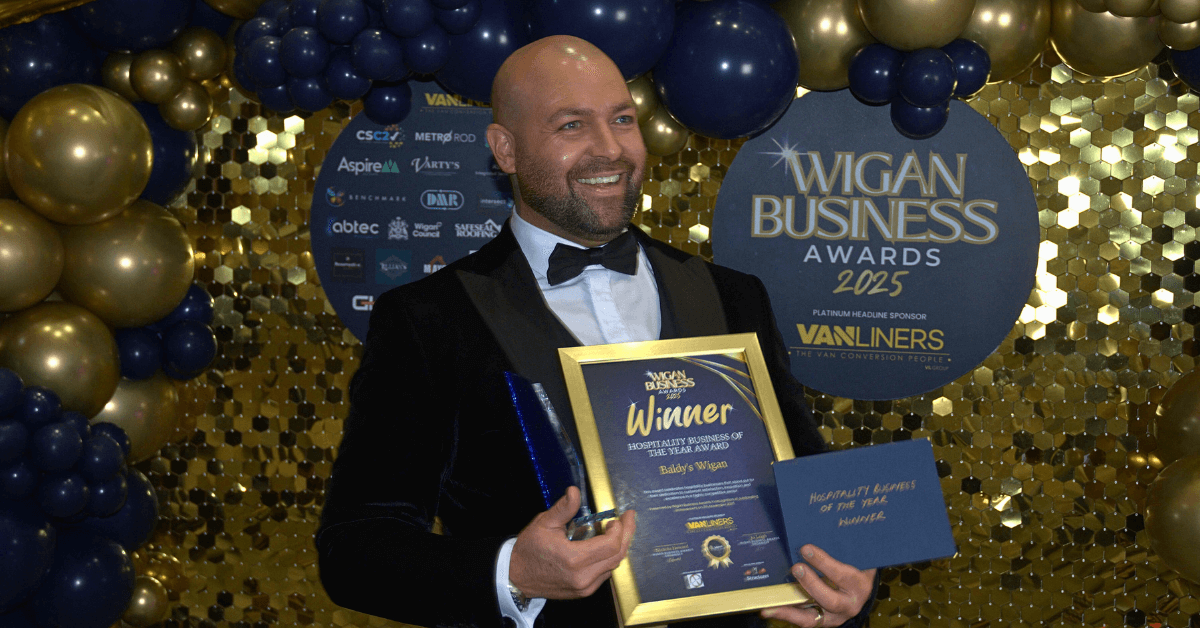Smiling awards winner in a tuxedo holding a framed certificate and trophy in front of the Wigan Business Awards 2025 backdrop with gold and navy balloons.