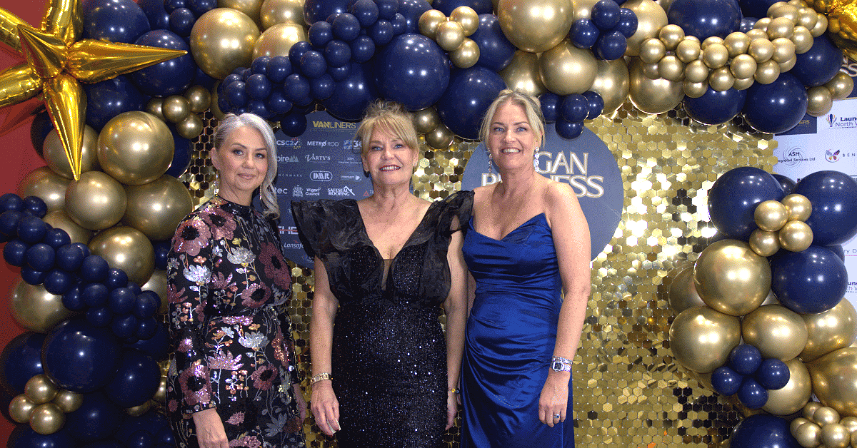 Three women pose in evening dresses in front of a Wigan Business Awards backdrop with gold sequin wall and navy and gold balloons.