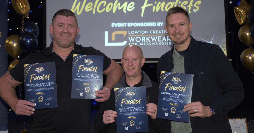 Three men smile at a Wigan Business Awards finalists event, holding finalist certificates in front of a Welcome Finalists screen.