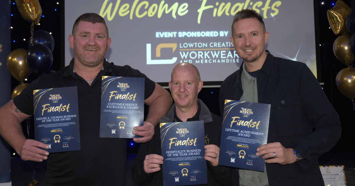 Three men smile at a Wigan Business Awards finalists event, holding finalist certificates in front of a Welcome Finalists screen.