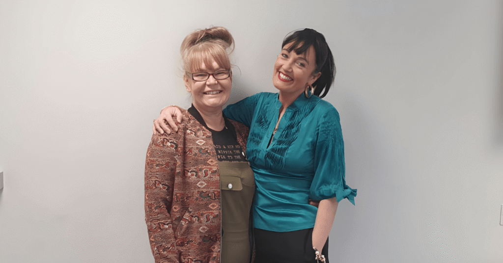 Two people stand against a plain wall. The person on the left wears glasses, a patterned jacket, and olive green overalls. The person on the right wears a teal blouse with ruffled details and black pants.