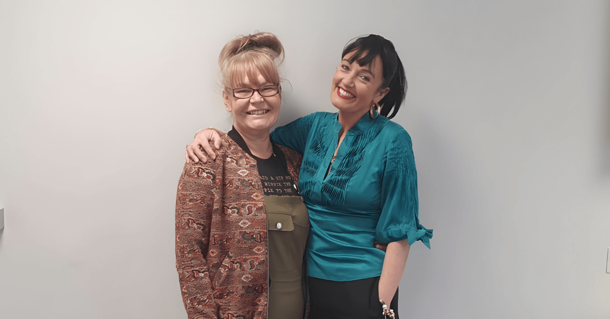 Two people stand against a plain wall. The person on the left wears glasses, a patterned jacket, and olive green overalls. The person on the right wears a teal blouse with ruffled details and black pants.