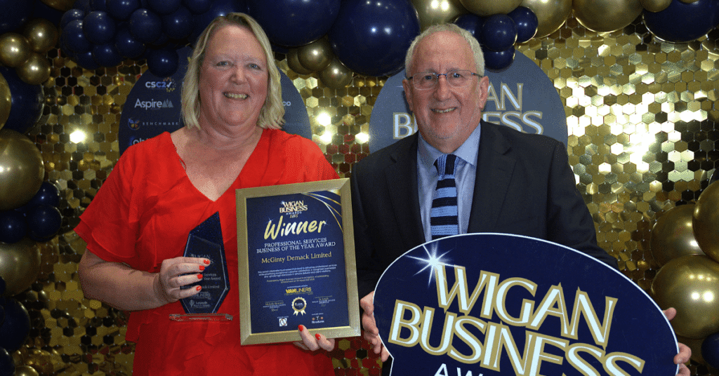 Two people are holding awards at the Wigan Business Awards. One person is holding a glass trophy and a framed certificate, while the other holds a large Wigan Business Awards winner sign. They are standing in front of a backdrop of gold and navy blue balloons.