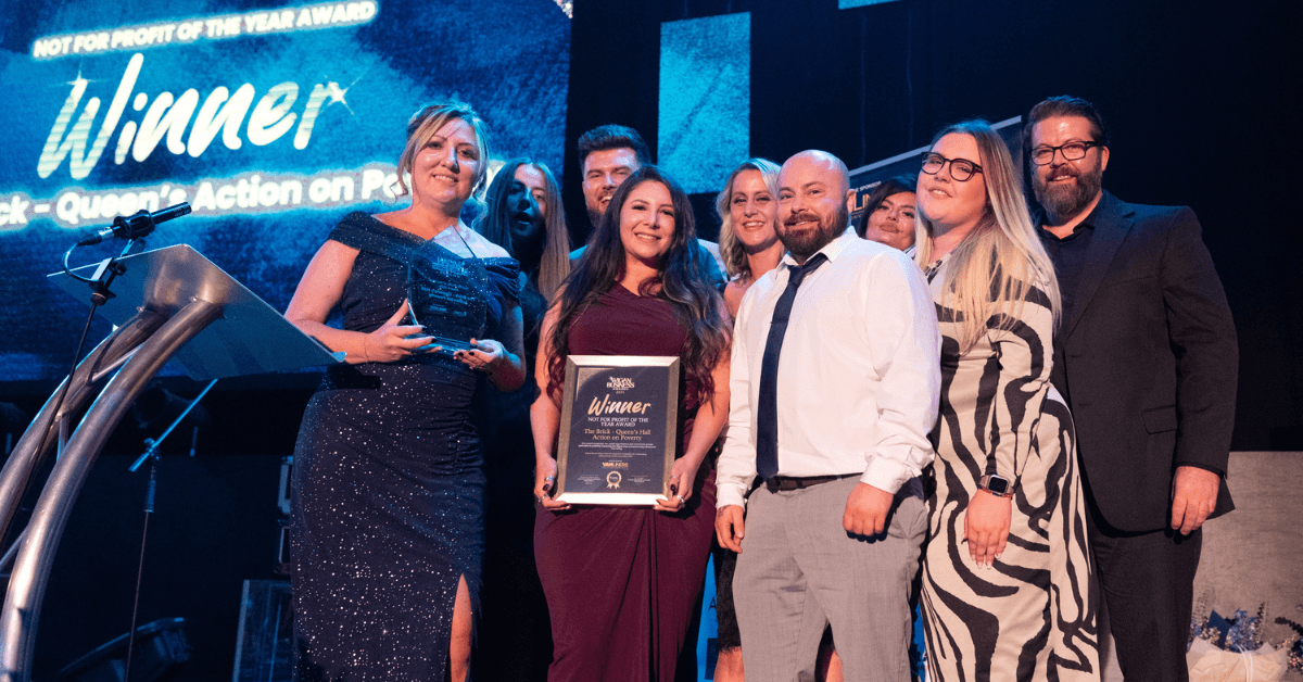 The Brick accepting an award. A woman in a sparkly blue dress holds a glass award, and another woman in a burgundy dress holds a framed certificate. The text on the screen behind them reads "Winner" and "NOT FOR PROFIT OF THE YEAR AWARD".