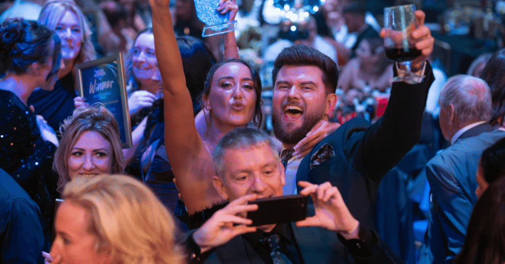 A group of people celebrating at Wigan Business awards ceremony. One person holds up a drink, another a trophy, and someone is taking a photo with their phone.