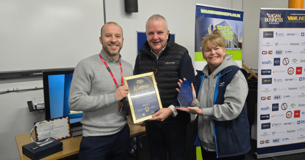 Three people holding a Wigan Business Awards winner certificate and trophy, standing in front of event banners at a business awards presentation.