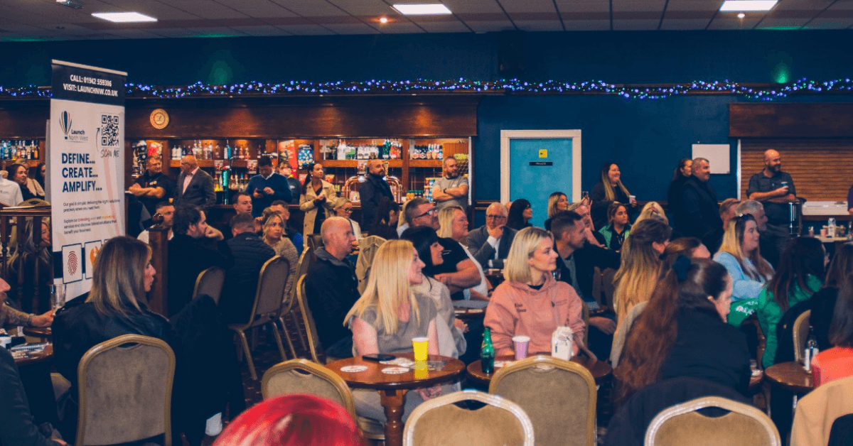 A large audience sits at round tables in a function room, watching a presentation off-camera. People are seated and standing around the space, with a bar at the back, a promotional banner on the left, and decorative string lights running along the wall. The room has a relaxed networking or business event atmosphere, with attendees listening attentively.