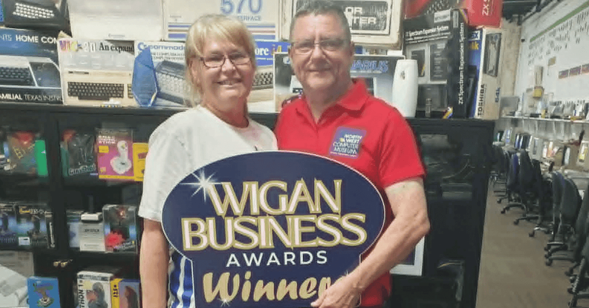 Two adults stand smiling in a computer museum, holding a large "Wigan Business Awards Winner" sign. Shelves behind them display vintage computers, keyboards, software, and technology memorabilia, with a row of computer workstations visible along one side of the room.