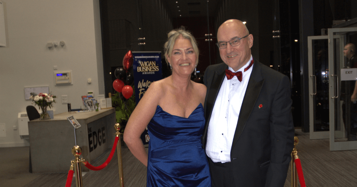 A man and woman pose together at a formal business awards event. The woman wears a dark blue evening dress, while the man wears a black tuxedo with a red bow tie. Behind them is signage for the Wigan Business Awards, with red balloons and a reception area visible.