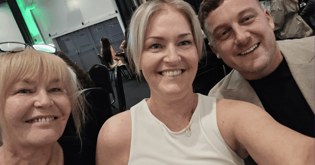 A group of three people smiling for a selfie at an indoor business event. Two women and one man are seated closely together, with event attendees and a conference setting visible in the background.