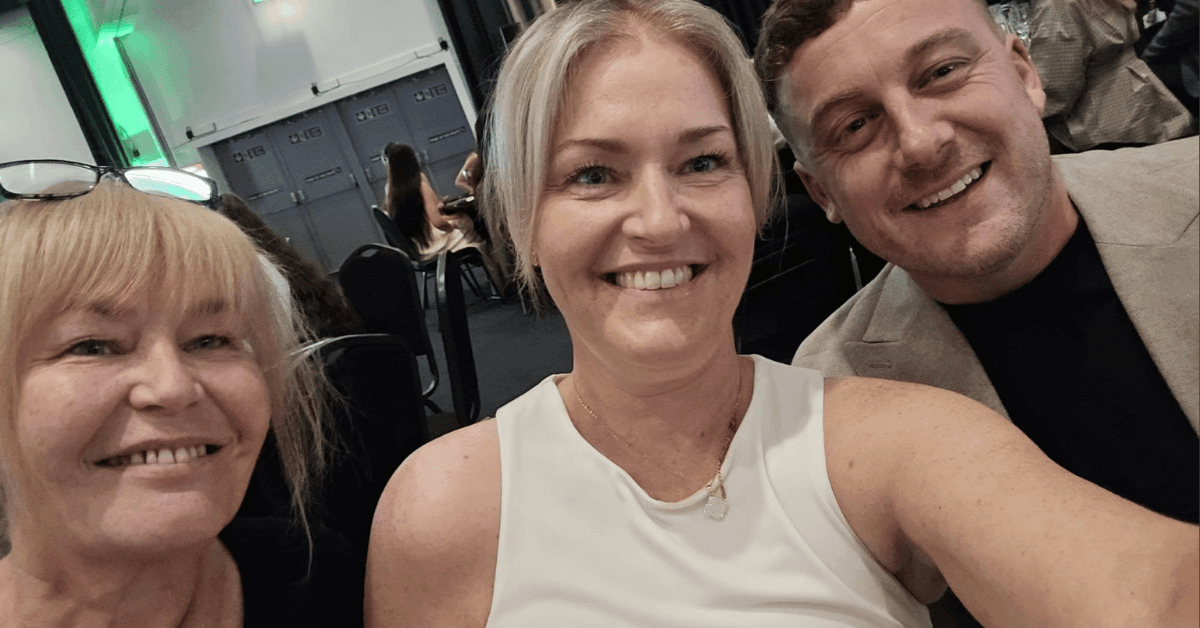 A group of three people smiling for a selfie at an indoor business event. Two women and one man are seated closely together, with event attendees and a conference setting visible in the background.