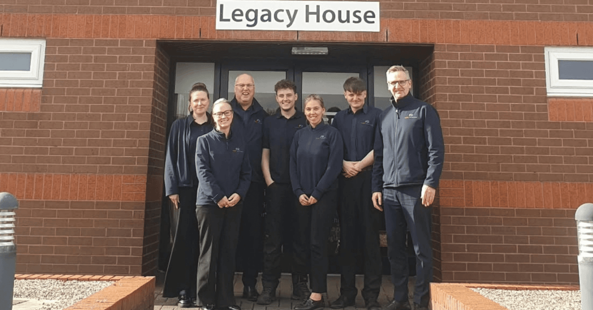 Closer group photo of eight Fleet Dynamic team members standing together outside the entrance to Legacy House, wearing branded navy workwear