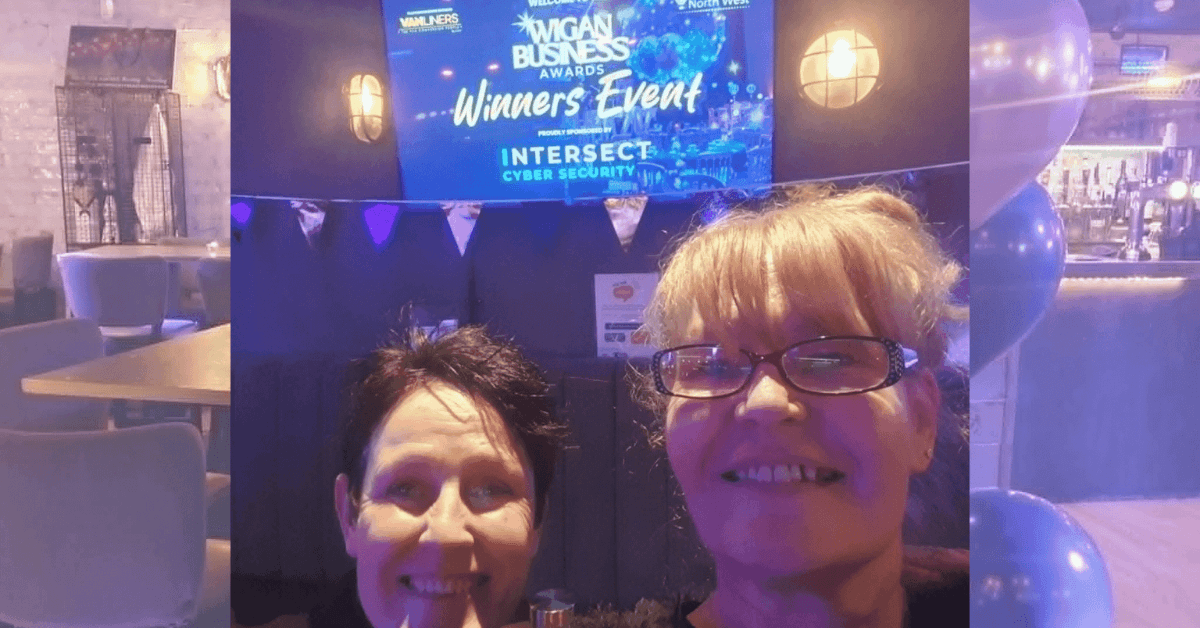 Two women smile for a selfie at the 2026 Wigan Business Awards Winners’ Event, sponsored by Intersect Cyber Security. The event venue features blue balloons, celebratory decorations and an illuminated awards sign.