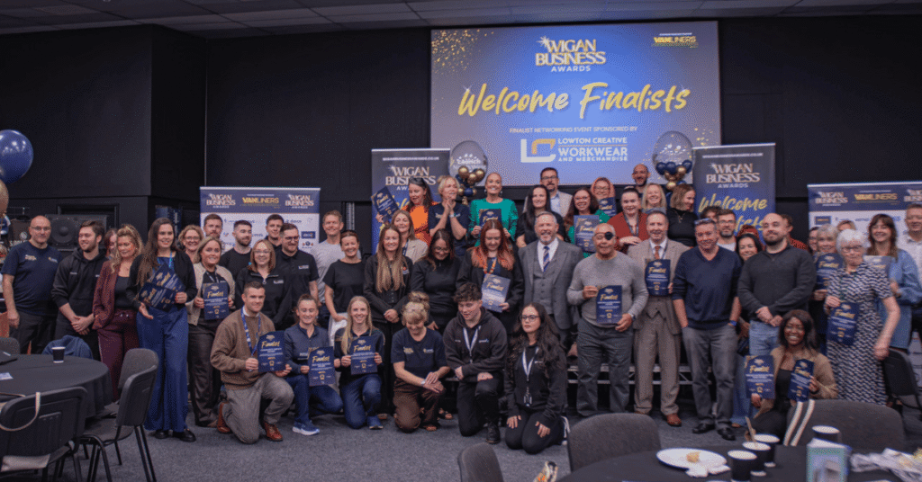 A large group of Wigan Business Awards 2026 finalists pose for a group photograph at a networking event. The group holds finalist certificates and awards in front of a stage backdrop reading “Welcome Finalists,” with blue and gold balloons, promotional banners and event signage.