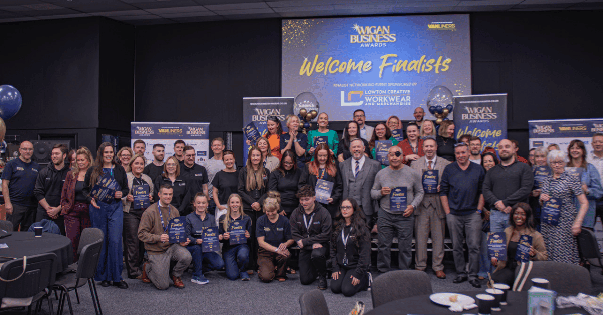 A large group of Wigan Business Awards 2026 finalists pose for a group photograph at a networking event. The group holds finalist certificates and awards in front of a stage backdrop reading “Welcome Finalists,” with blue and gold balloons, promotional banners and event signage.