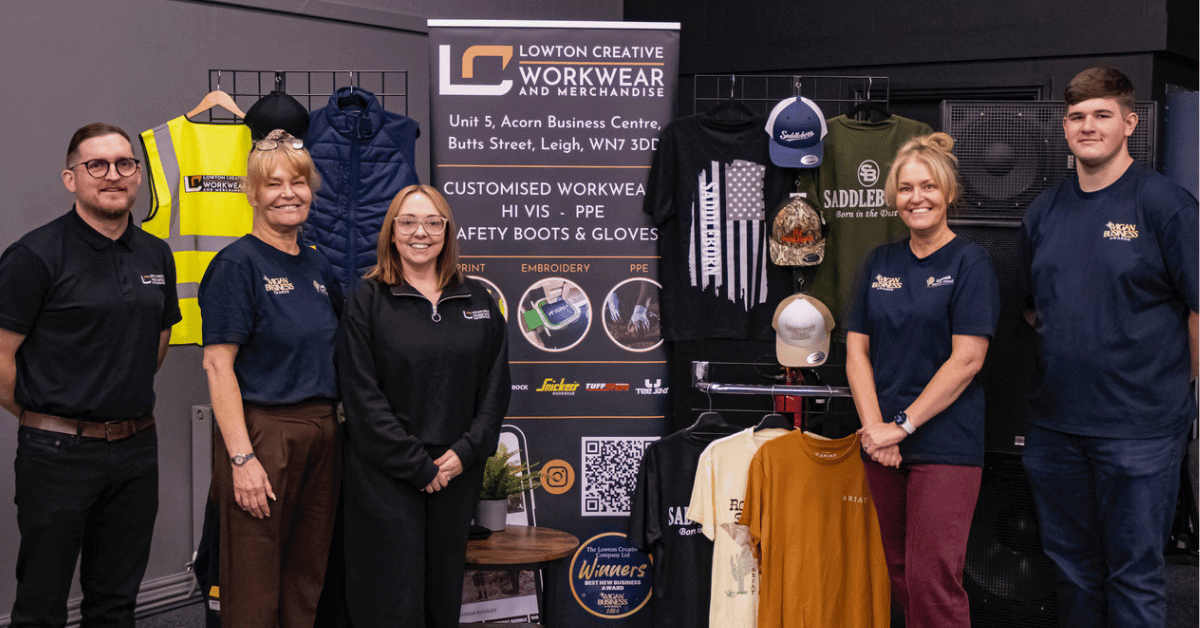 Five members of the Lowton Creative Workwear and Merchandise team pose at the Wigan Business Awards finalists’ networking event. Behind them is a display of branded workwear, including high-visibility jackets, polo shirts, hoodies, caps and other merchandise, alongside a promotional banner advertising customised workwear, PPE, embroidery and printing services.
